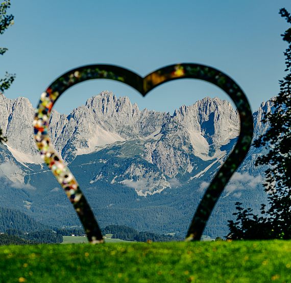 Heart-shaped sculpture frames a picturesque mountain range with lush green foreground and clear blue sky.