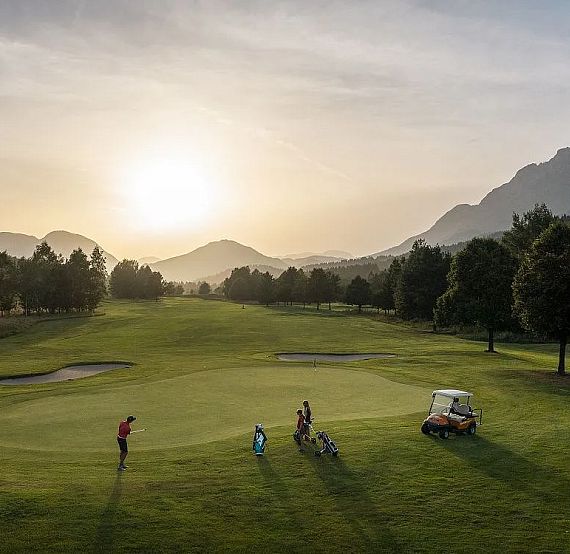 Golfers on a lush, green course at sunset, with mountains in the distance under a colorful sky.
