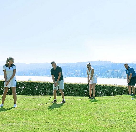 Eine Gruppe von fünf Personen steht auf einem sonnigen Golfplatz mit Blick auf einen See und bewaldete Hügel. Die Menschen bereiten sich auf einen Schlag vor.