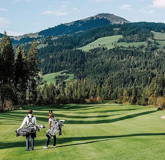 Two golfers carrying their bags on a green course surrounded by mountains and forests on a sunny day. The landscape is lush and picturesque.
