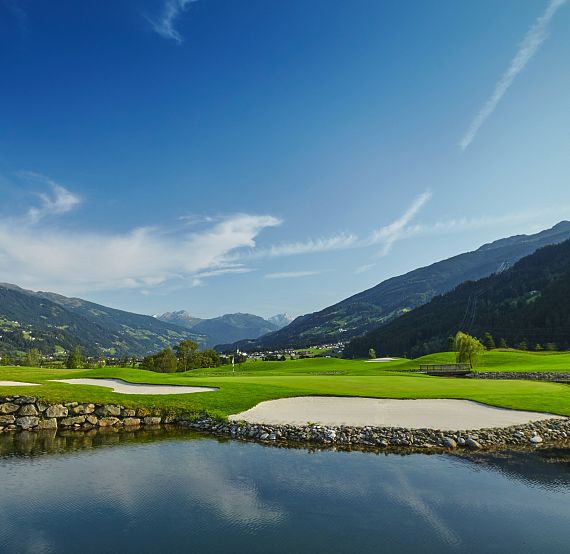 Grüner Golfplatz mit Sandbunkern, umgeben von Hügeln und blauem Himmel. Ein ruhiger Teich im Vordergrund spiegelt die idyllische Landschaft wider.