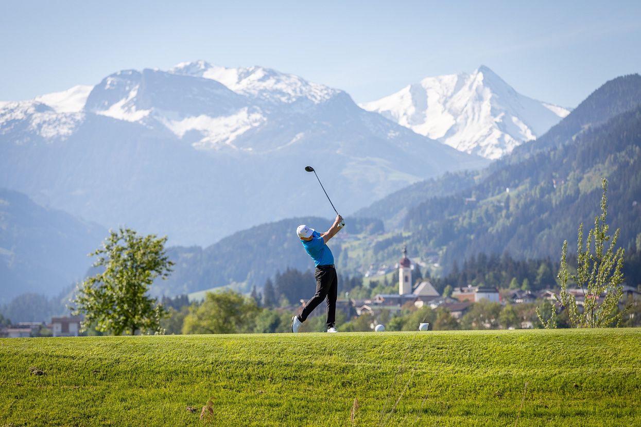 Über eine grüne Golfwiese schwingt ein Golfer seinen Schläger, umgeben von majestätischen, schneebedeckten Bergen unter einem klaren blauen Himmel.