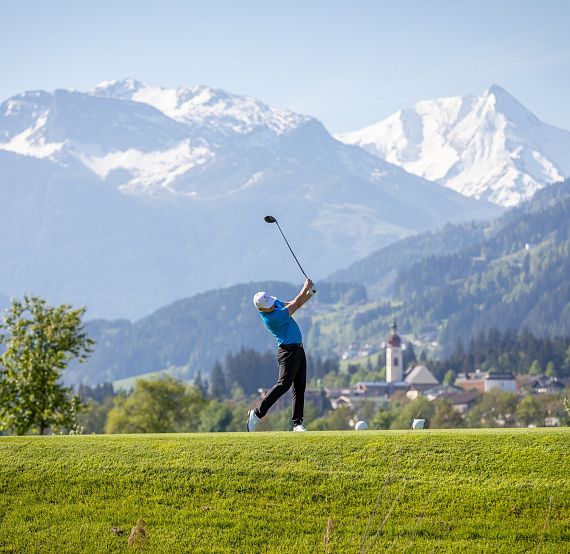 Über eine grüne Golfwiese schwingt ein Golfer seinen Schläger, umgeben von majestätischen, schneebedeckten Bergen unter einem klaren blauen Himmel.