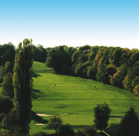 Eine weitläufige Golfanlage umgeben von üppigem, grünem Wald unter einem klaren blauen Himmel. Im Vordergrund sind gepflegte Rasenflächen und Bäume sichtbar.