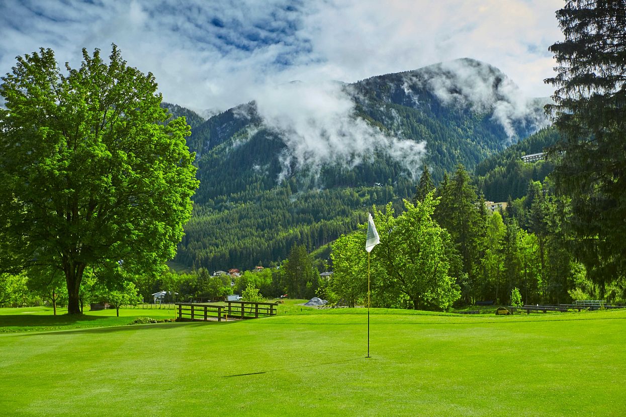Grüne Golflandschaft mit einem Fahnenstock auf dem Grün, umgeben von dichten Bäumen und im Hintergrund eindrucksvollen Bergen unter bewölktem Himmel.