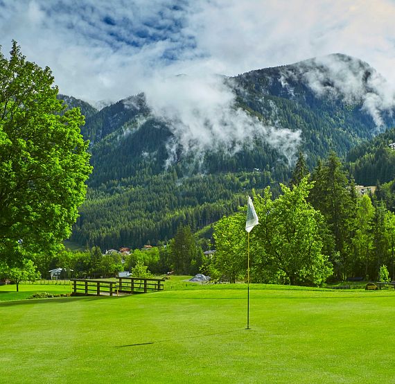 Grüne Golflandschaft mit einem Fahnenstock auf dem Grün, umgeben von dichten Bäumen und im Hintergrund eindrucksvollen Bergen unter bewölktem Himmel.