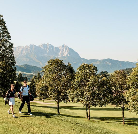 Zwei Personen gehen mit Golftaschen über einen grünen Golfplatz, im Hintergrund erheben sich majestätische Berge und Bäume stehen in der Nähe.