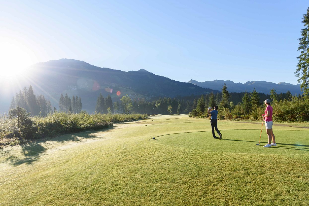 Zwei Golfer auf einem weitläufigen grünen Golfplatz in den Bergen, bei strahlendem Sonnenschein. Einer schlägt gerade ab, während der andere zusieht.