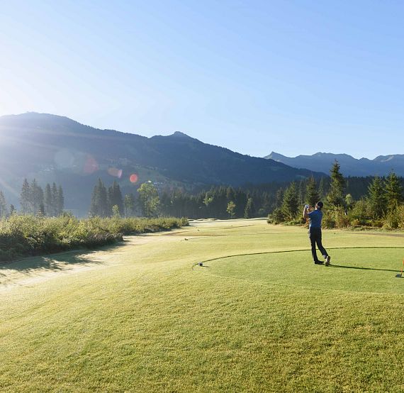 Zwei Golfer auf einem weitläufigen grünen Golfplatz in den Bergen, bei strahlendem Sonnenschein. Einer schlägt gerade ab, während der andere zusieht.