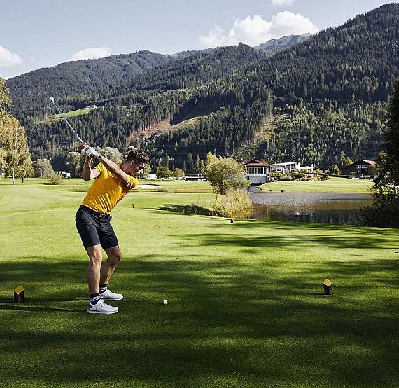 Golfer in yellow shirt swings on a green course with mountains in the background, under a blue sky.