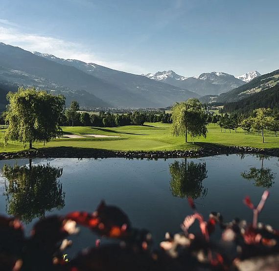 A picturesque golf course with green fairways, a pond, and mountains in the background under a clear blue sky.