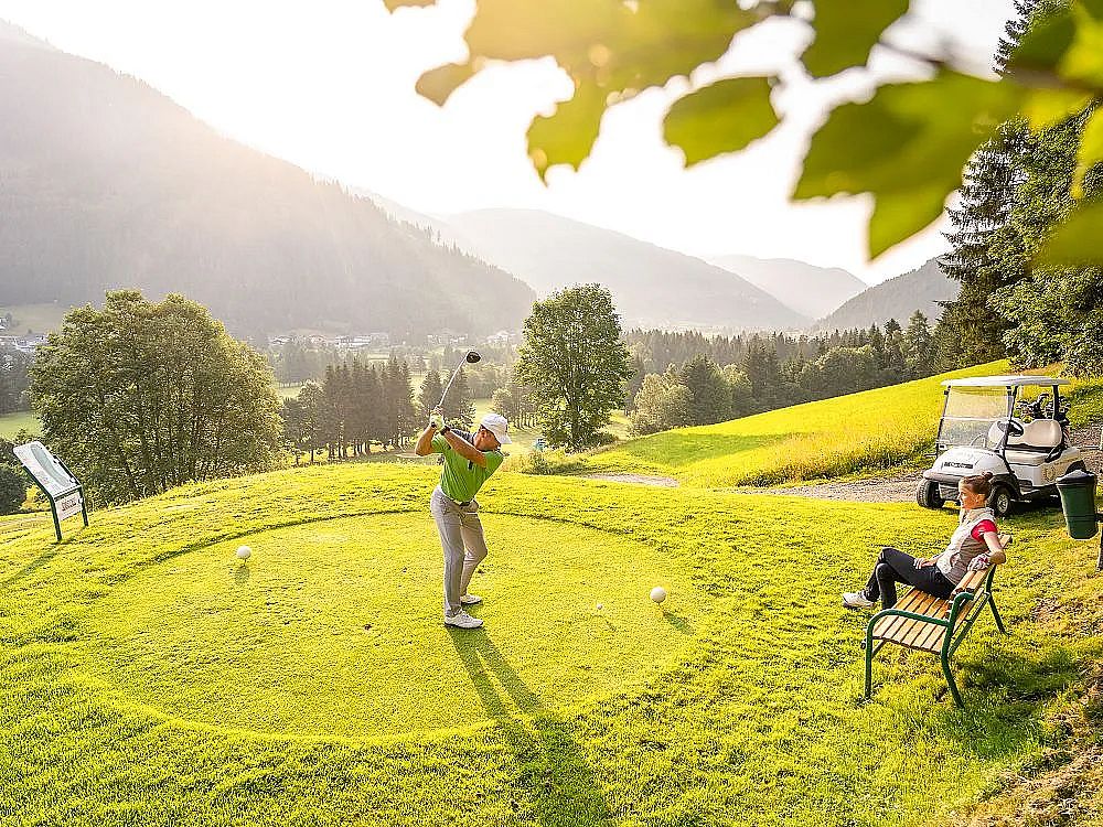 Golfer schlägt auf einem sonnigen, grünen Golfplatz ab, während eine Person auf einer Bank sitzt und zusieht. Im Hintergrund sind Bäume und Berge zu sehen.