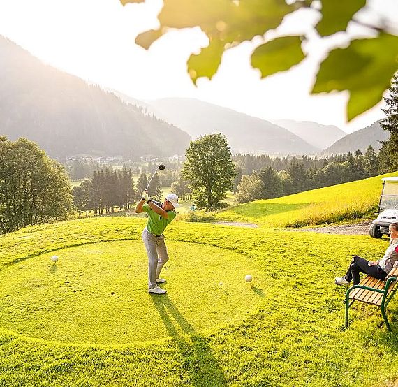 A golfer swings on a green golf course surrounded by tree-covered mountains. Another person sits on a bench nearby, watching the game under a sunlit sky.