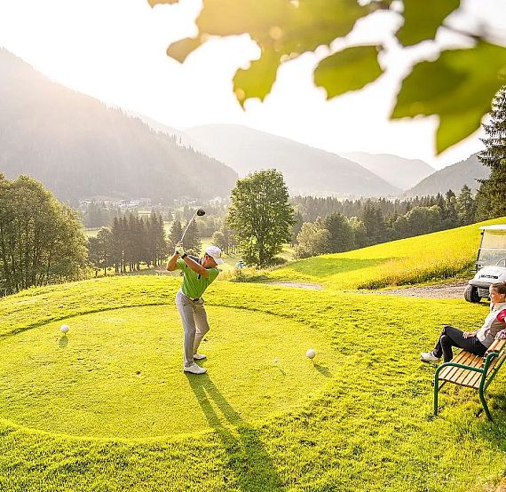 Golfer tees off on a green course surrounded by mountains and trees, under a bright blue sky. A person sits nearby, watching from the side. Sunlight filters through leaves.