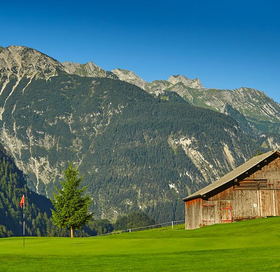 A wooden cabin sits on a green meadow with towering mountains in the background under clear blue skies.