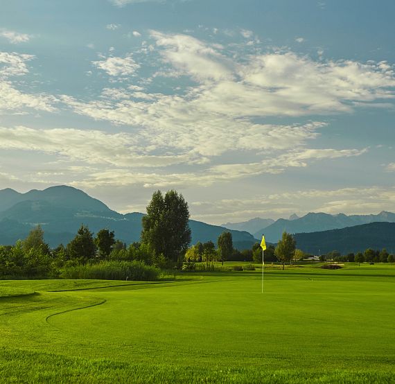 A lush green golf course with a flag, surrounded by mountains and a blue sky with scattered clouds, creating a serene natural setting.