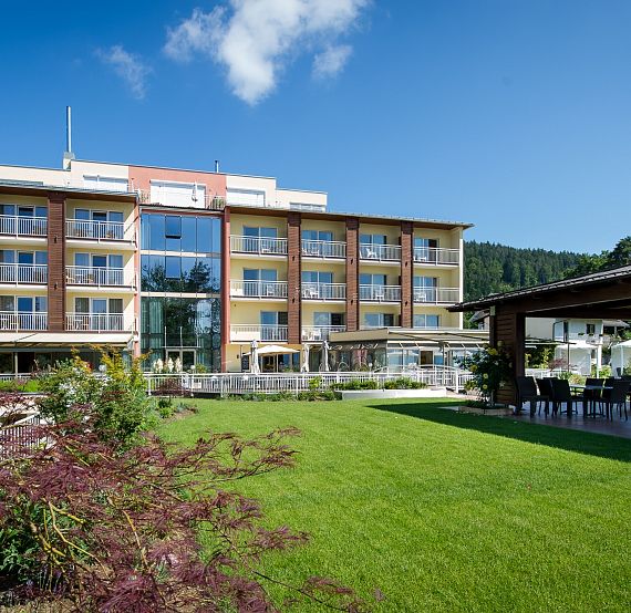 Modernes Hotelgebäude mit Glasfassade, umgeben von gepflegtem Garten und Terrasse. Liegt in einer grünen, sonnigen Gegend mit leicht bewölktem Himmel.