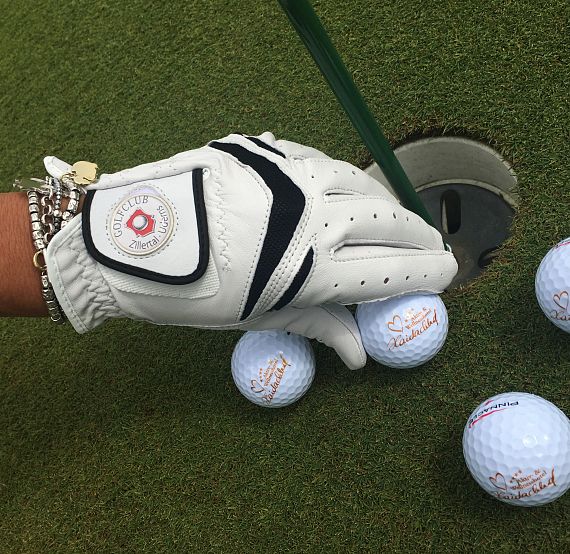 A gloved hand picks up a golf ball from a hole on a lush green surface, with other balls around, indicating a practice or successful putting session.