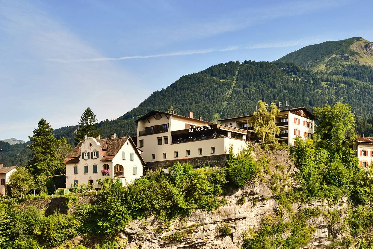 A village with houses on a cliff, surrounded by green mountains and under a blue sky. The buildings and natural scenery create a serene landscape.
