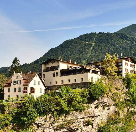 Ein malerisches Dorf auf einem Hügel mit traditionellen Häusern und üppigem Grün umgeben, vor einer Kulisse von Bergen und klarem Himmel.