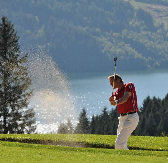 A golfer in red shirt and white pants swings in a sand trap, surrounded by lush greenery and a shimmering lake in the background.