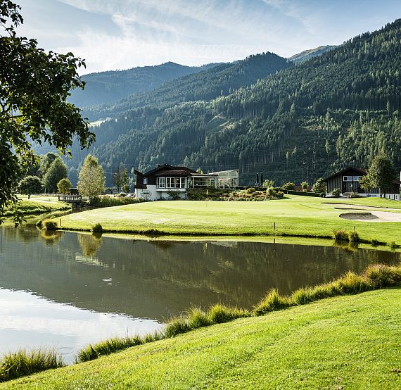 A golf course by a serene lake, surrounded by lush green hills and trees, with mountains in the background under a blue sky.