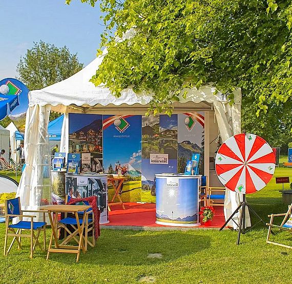 Outdoor booth with tent, display stands, a spinning game wheel, and seating on grass under trees in a park setting.