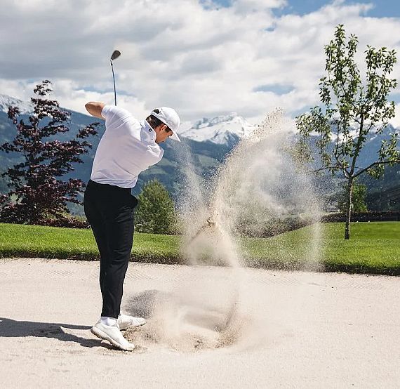 A golfer swings in a sandy bunker, with lush greenery and snowy mountains under a blue sky in the background.