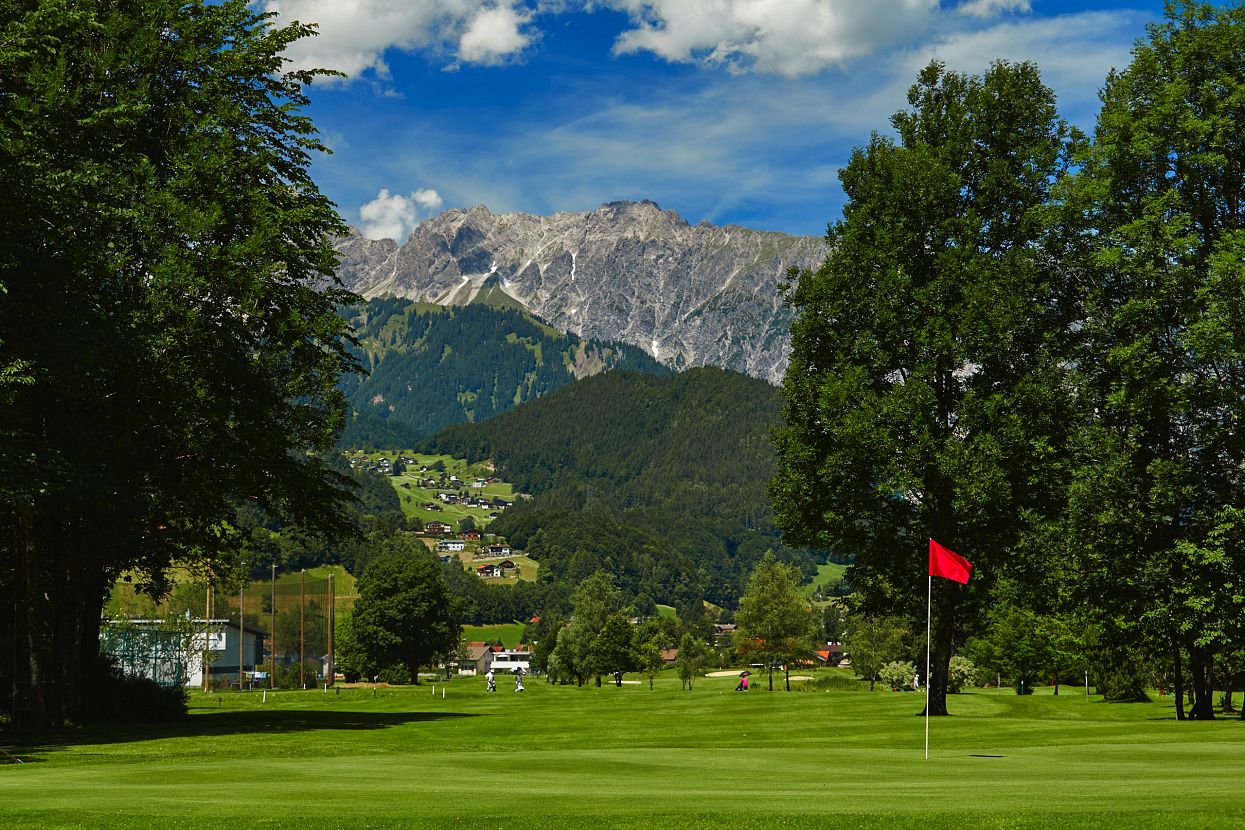 Eine grüne Golfanlage mit roter Flagge im Vordergrund, umgeben von hohen Bäumen. Im Hintergrund sind majestätische Berge unter einem blauen Himmel zu sehen.
