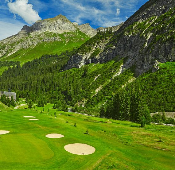 Berglandschaft mit einem grünen Golfplatz im Vordergrund, umgeben von Nadelbäumen. Im Hintergrund erheben sich imposante Berge unter einem blauen Himmel.