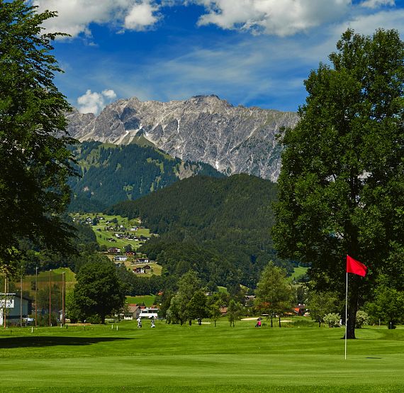 Eine grüne Golfanlage mit roter Flagge im Vordergrund, umgeben von hohen Bäumen. Im Hintergrund sind majestätische Berge unter einem blauen Himmel zu sehen.