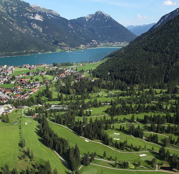 Luftaufnahme einer malerischen Berglandschaft mit einem See. Ein Dorf mit grünen Wiesen und ein gut gepflegter Golfplatz sind zu erkennen.