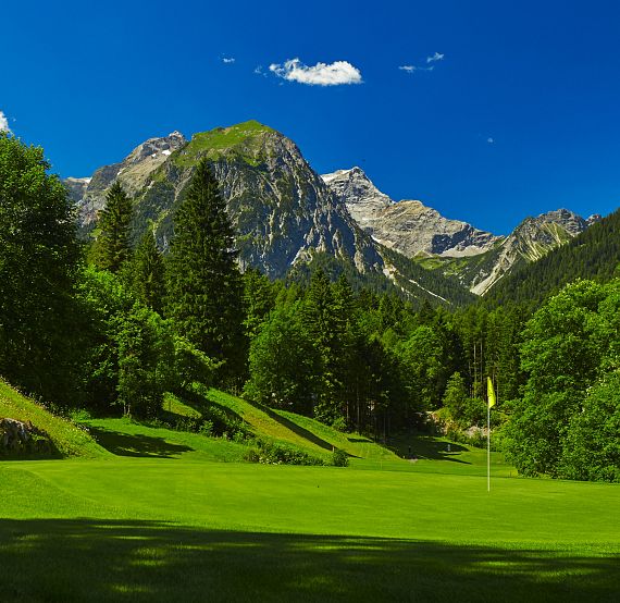 Grüne Golfbahn umgeben von dichtem Wald, dahinter eindrucksvolle Bergkulisse unter strahlend blauem Himmel. Eine Fahne markiert das Loch auf dem Grün.
