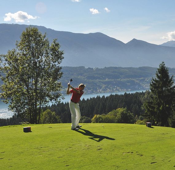 Golfer swings on a green course with mountains and a lake in the background under a clear blue sky, surrounded by trees.