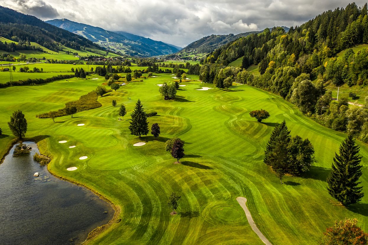 Luftbild eines weitläufigen Golfplatzes in einer hügeligen Landschaft mit saftigem Grün, Sandbunkern und einem kleinen See. Bewaldete Hügel im Hintergrund.