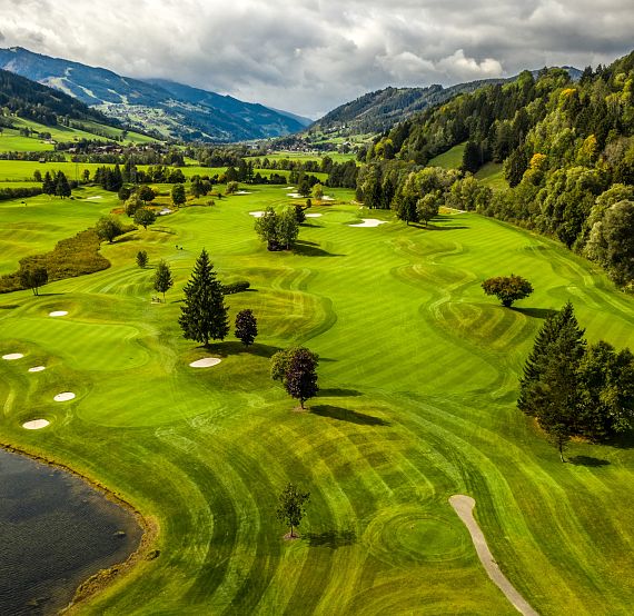 Luftbild eines weitläufigen Golfplatzes in einer hügeligen Landschaft mit saftigem Grün, Sandbunkern und einem kleinen See. Bewaldete Hügel im Hintergrund.