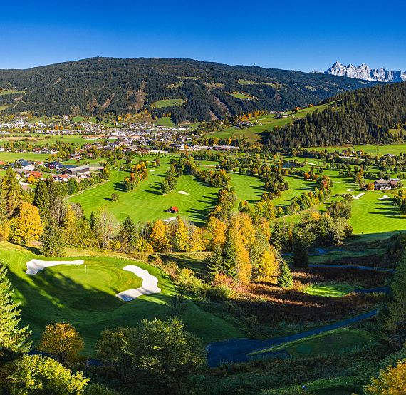 A scenic view of a vast golf course with rolling hills and mountains in the distance, under a clear blue sky. Trees display vibrant autumn foliage.