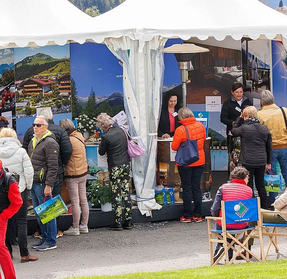 People visiting booths at an outdoor fair. Some are seated while others stand and interact with exhibitors. Bright tents and lively atmosphere visible.