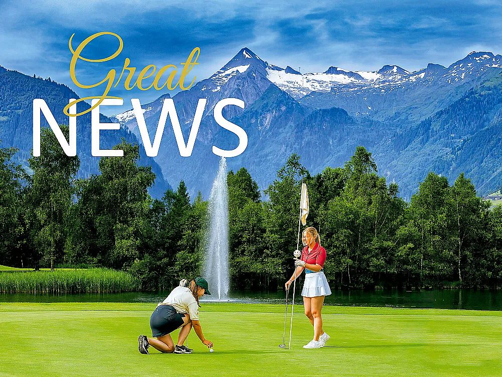 Two golfers on a green, one bending and another holding a flag. Mountains and a fountain in the scenic background under a blue sky.