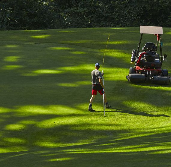 Ein Mann in sportlicher Kleidung steht auf einem Golfplatz. Neben ihm ist ein Rasenmäherfahrzeug. Die Sonne wirft Schatten auf das grüne Gras.