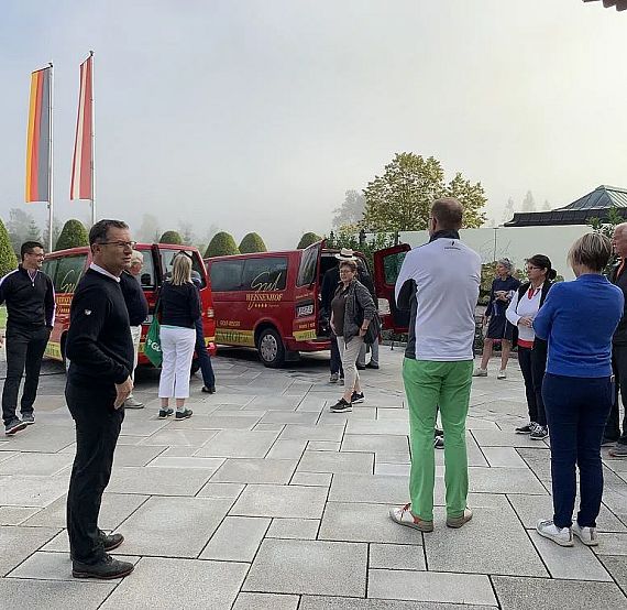 Eine Gruppe von Menschen steht auf einem gepflasterten Platz vor roten Fahrzeugen, mit mehreren Landesflaggen im Hintergrund an Fahnenmasten.