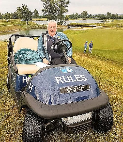 A man sits in a golf cart labeled "Rules" on a grassy golf course with two other people in the distance.
