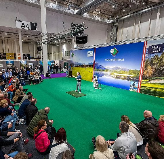Crowd at an indoor golf event with a stage and promotional banners. People watch presentations or demonstrations on a putting green area.