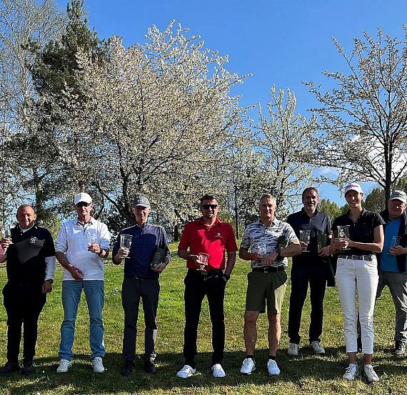 A group of people stands in a park holding trophies, smiling. Trees with blossoms are in the background under a clear, blue sky.