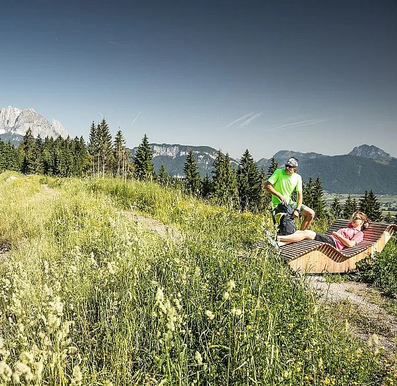 A scenic outdoor setting with a person relaxing on a reclined seat in a grassy meadow. Mountains and a clear sky are visible in the background.