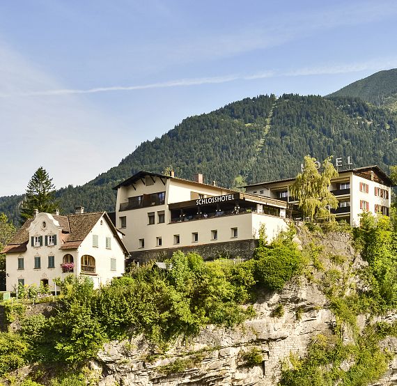 Scenic view of a hotel nestled on a hillside, with forested mountains and a clear sky in the background, surrounded by green foliage. Tranquil and picturesque setting.