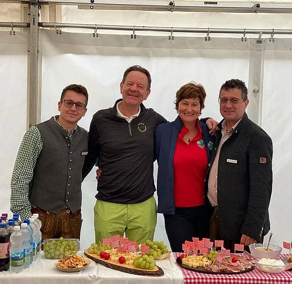 Vier Personen stehen lächelnd hinter einem Tisch mit Snacks und Getränken in einem Festzelt. Der Tisch ist mit rot-weiß kariertem Stoff bedeckt.