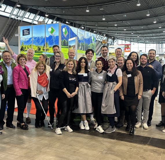 Eine Gruppe von Menschen posiert lächelnd für ein Foto auf einer Messe. Im Hintergrund sind bunte Stände und Banner mit Landschaftsmotiven zu sehen.