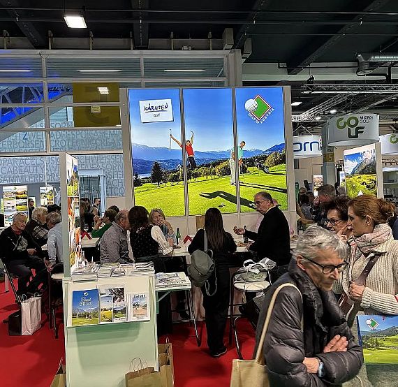 Trade show booth with a golf course backdrop, crowded with people interacting and discussing. Displays and literature are visible around the booth.