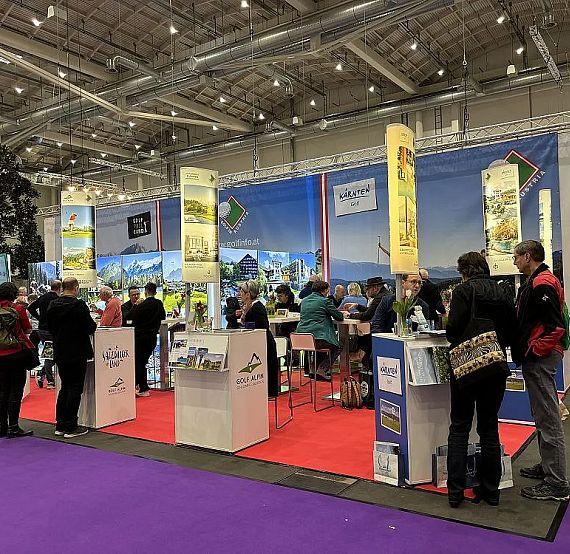 Busy trade show booth with people networking, colorful banners, and brochures, suggesting a promotional setup in a large exhibition hall.